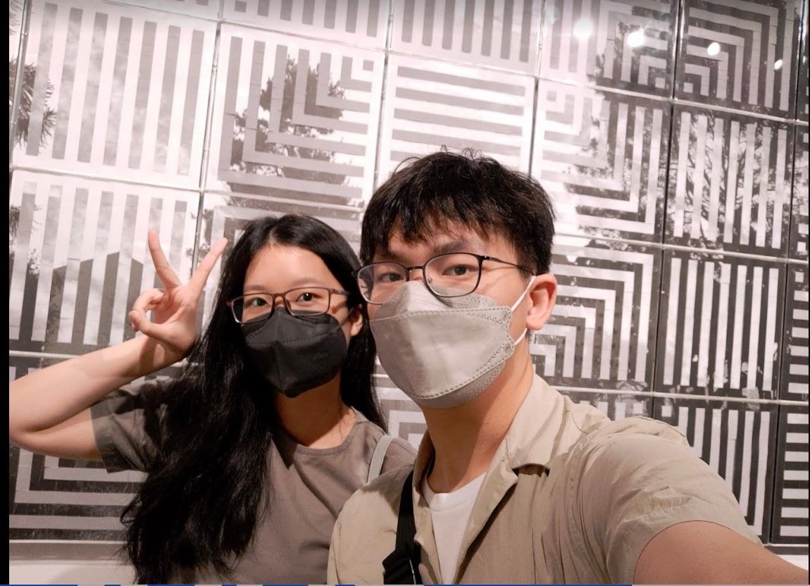 Rey and Feli wearing masks and taking a selfie in front of a patterned wall while Feli makes a peace sign