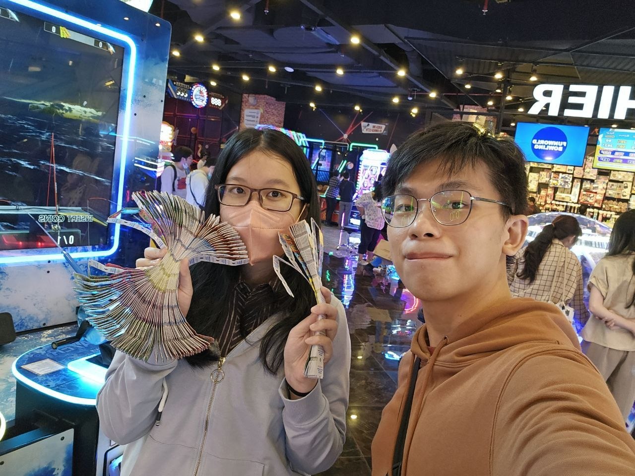 Rey and Feli at an arcade, smiling for a selfie while Feli holds a fan-shaped stack of tickets